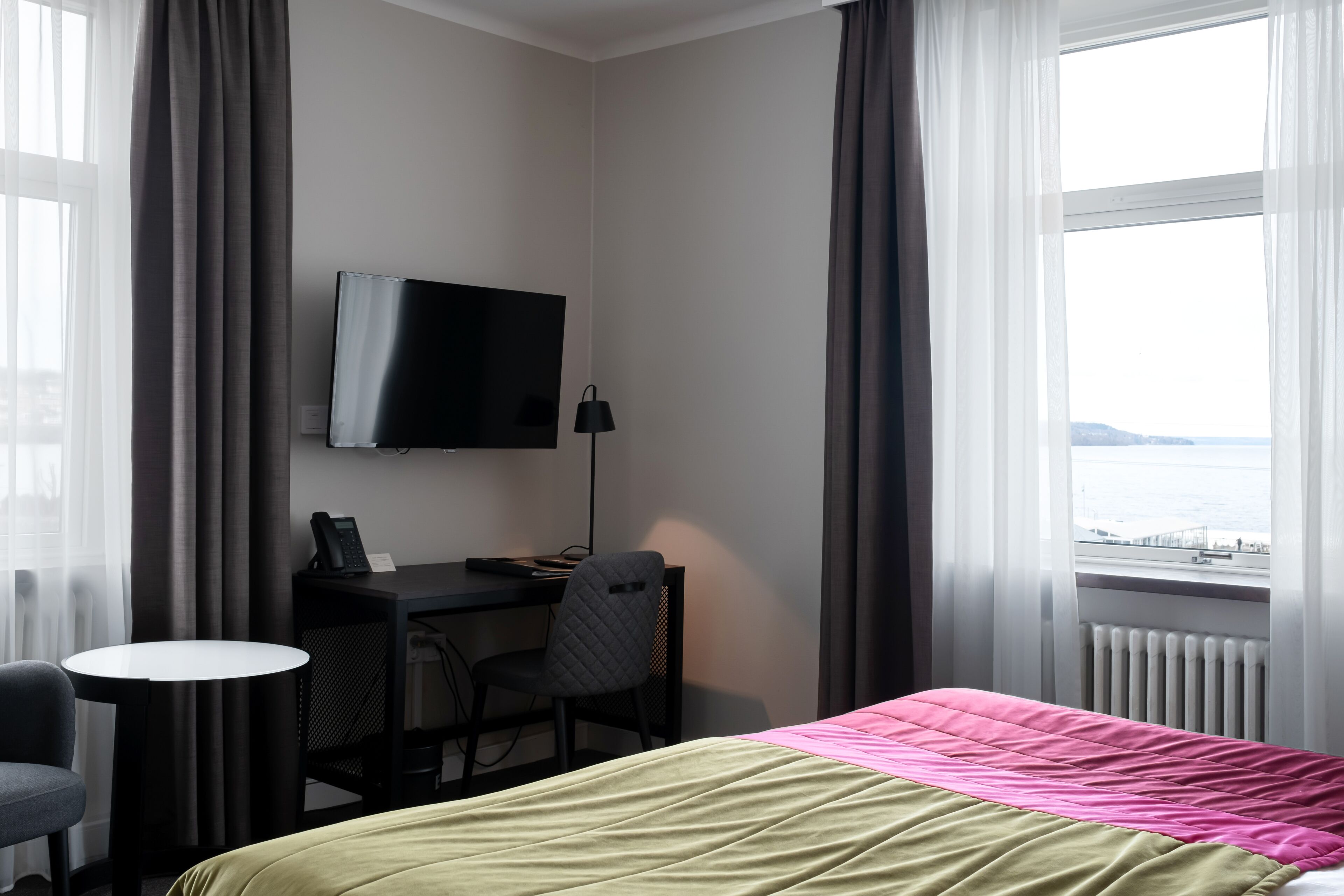 A hotel room with a bed, a desk with a chair, a television, and a view of the sea.