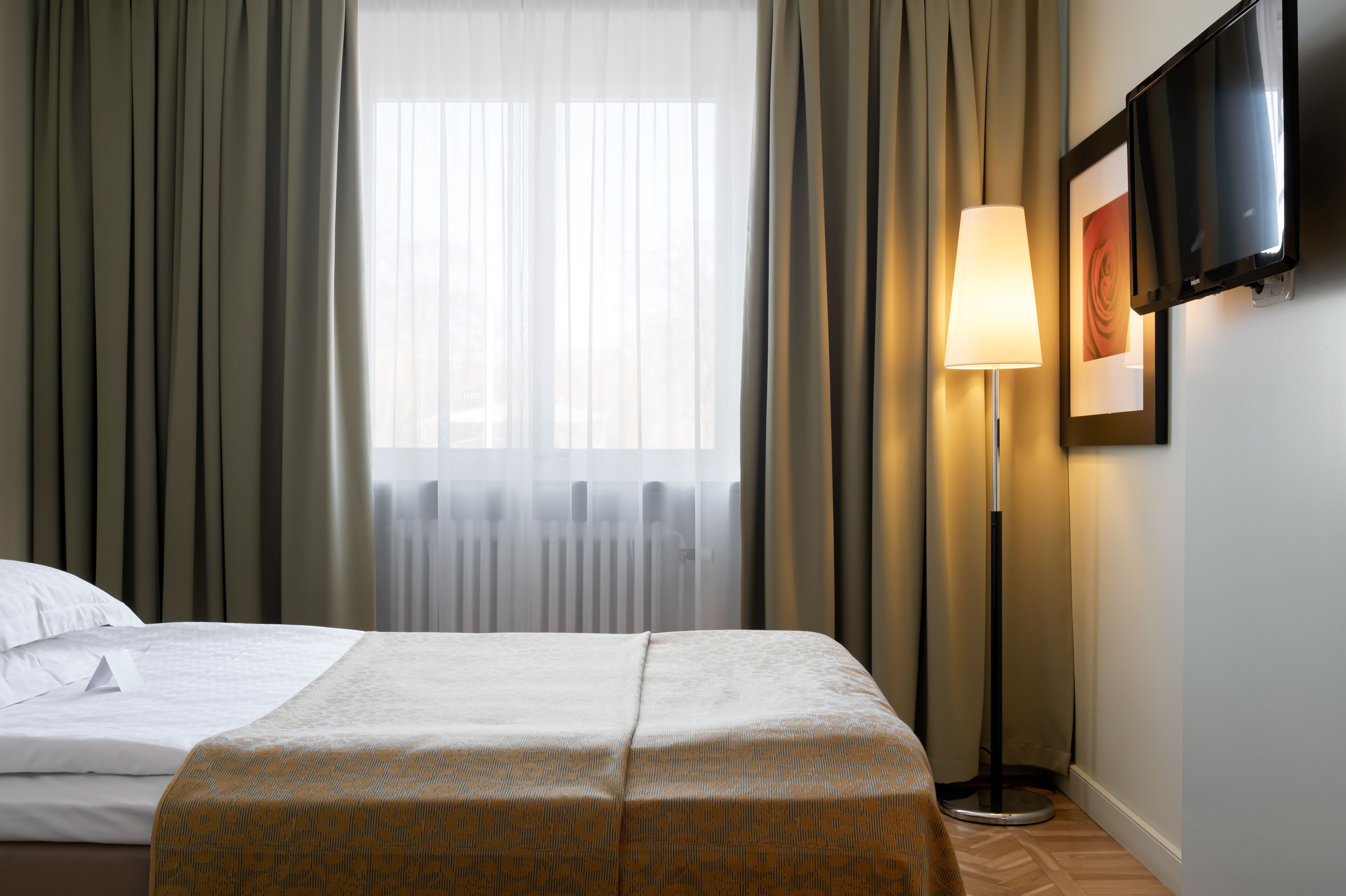 A hotel room with a bed, a standing lamp, a TV, and a window with curtains.