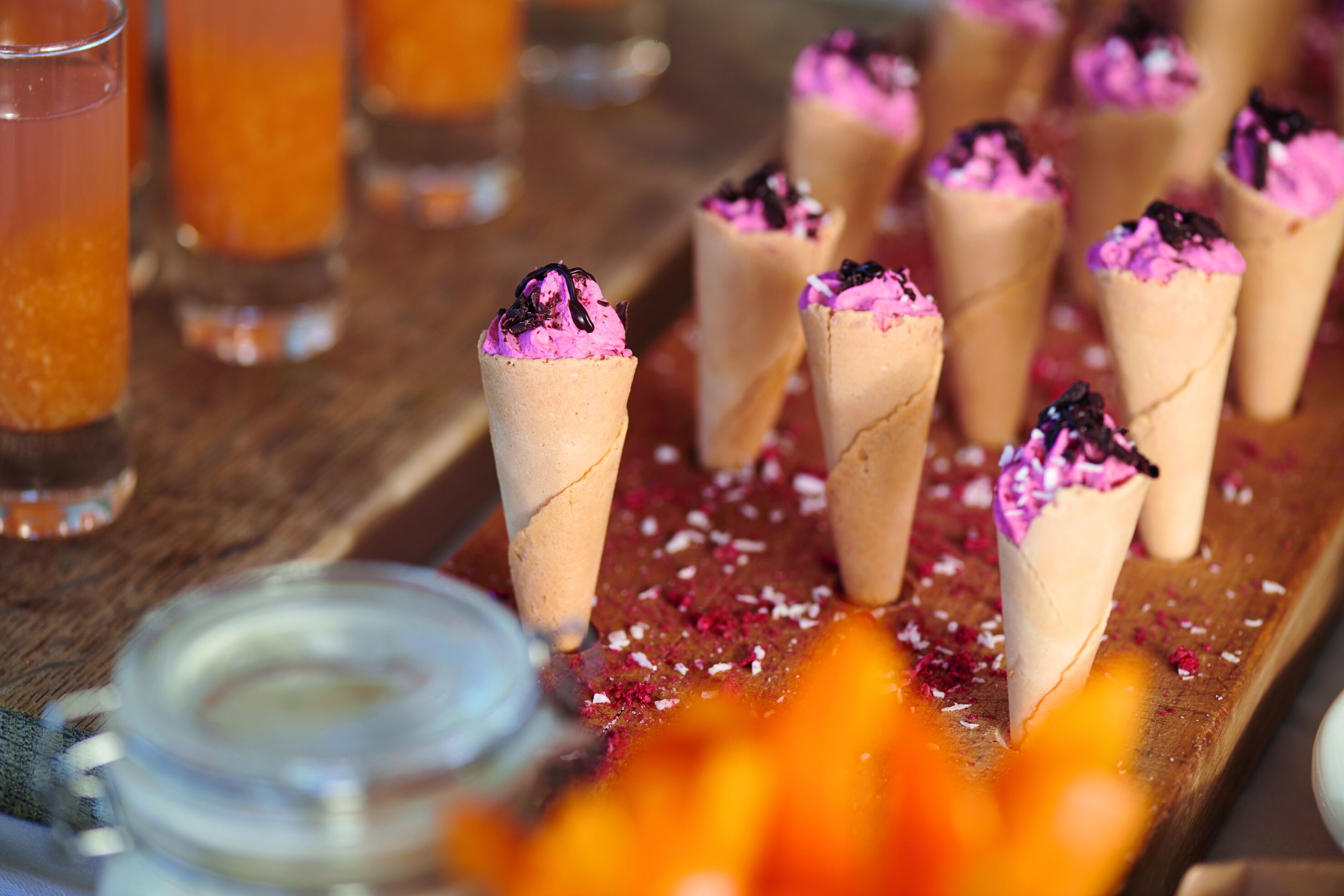 Una mesa de madera con varios conos de helado coronados con ingredientes rosas y negros, y un vaso de bebida de naranja.