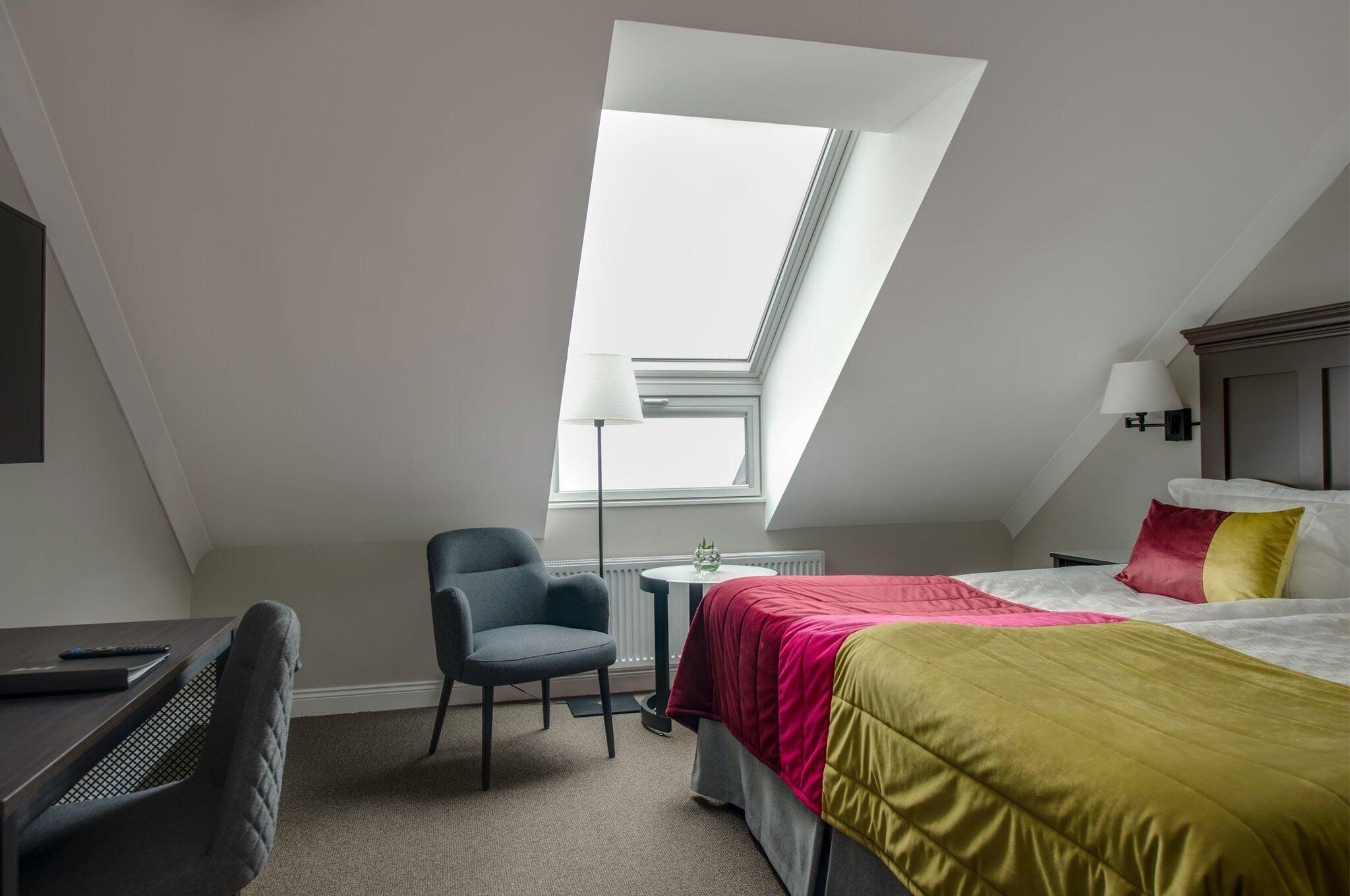 A small, modern room with a sloped ceiling, a bed with colorful bedding, a desk with a chair, and a small table with a lamp.