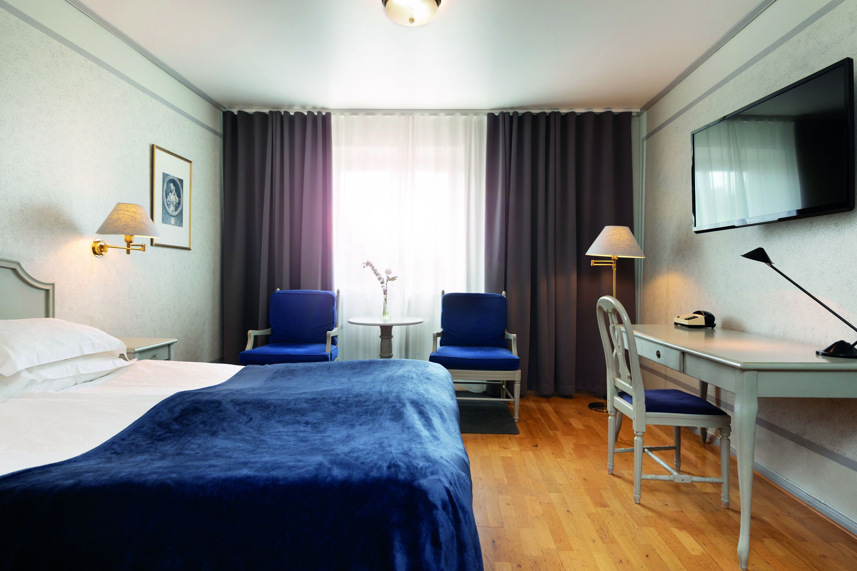 A hotel room with a bed, two chairs, a desk, a TV, and a window with curtains.