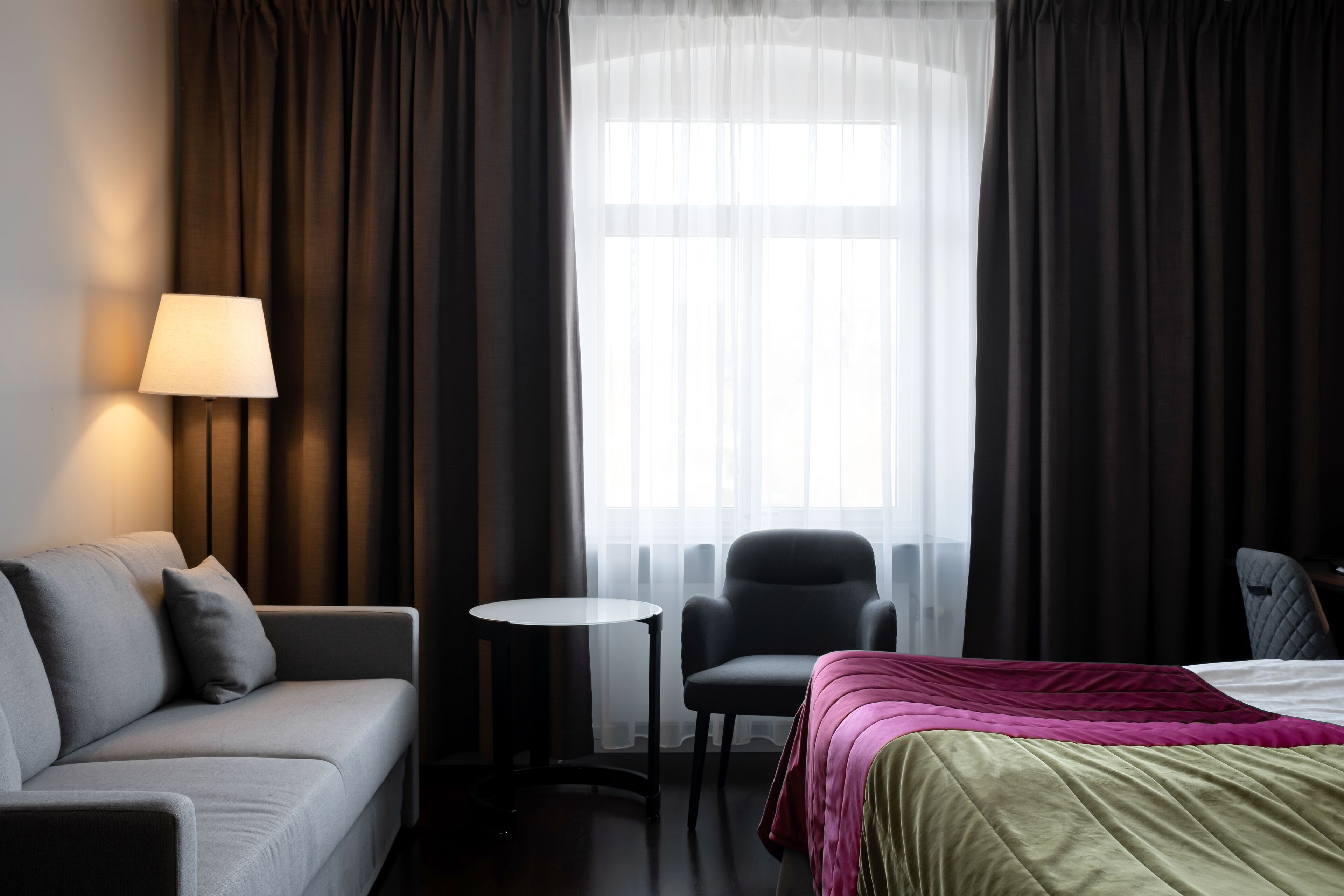 Una habitación de hotel con cama, sofá, sillón, una mesita y una lámpara.