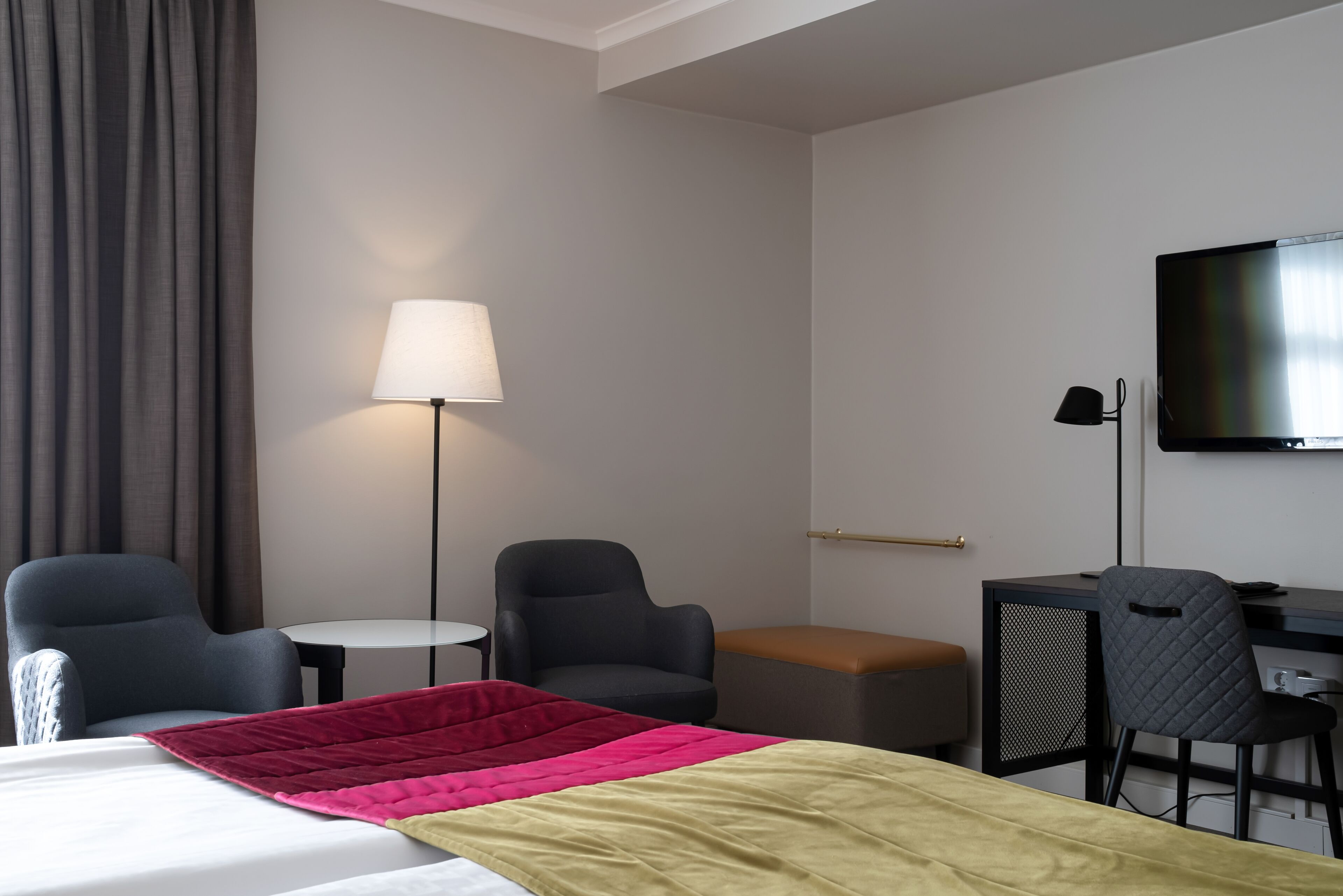 A hotel room with a bed, two armchairs, a desk, a television, and a lamp.