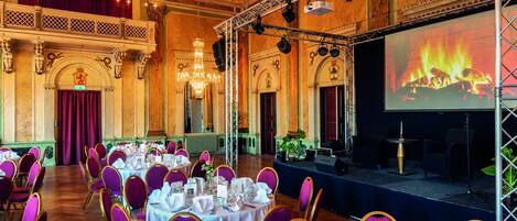 A banquet hall with round tables set for a formal event, a stage with a large screen displaying a fire, and ornate chandeliers.
