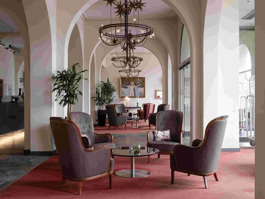 Un vestíbulo amplio con techos altos, columnas de arco y un candelabro central. Hay varios sillones y una mesa redonda con un florero.