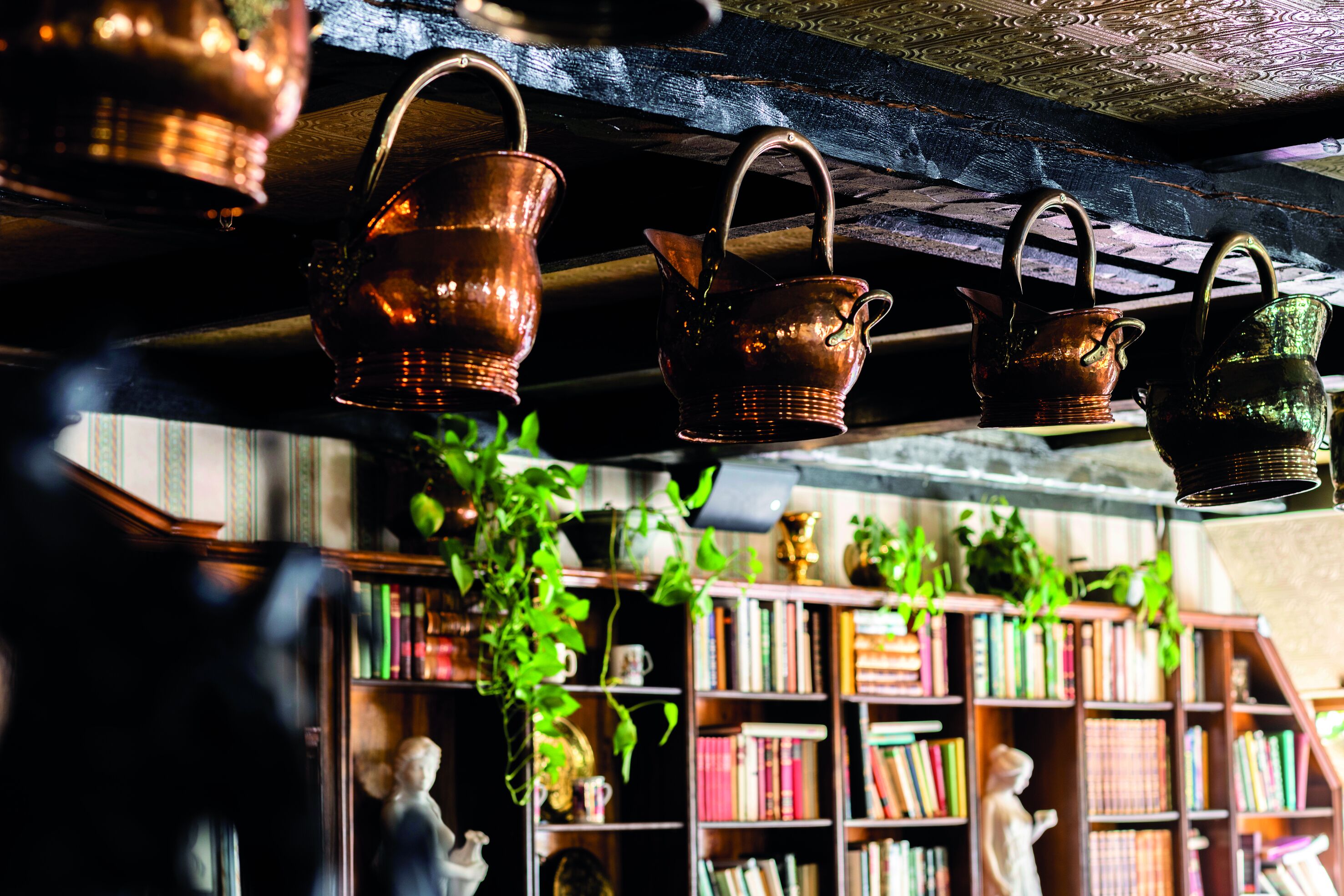 A rustic interior with copper pots hanging from the ceiling, a bookshelf filled with books, and a statue.