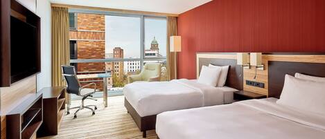 A hotel room with two beds, a desk, a chair, a TV, and a large window with a city view.