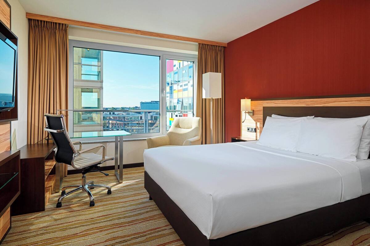 Habitación de hotel con una cama grande, un escritorio con una silla, un televisor y vistas a la ciudad a través de un ventanal amplio.
