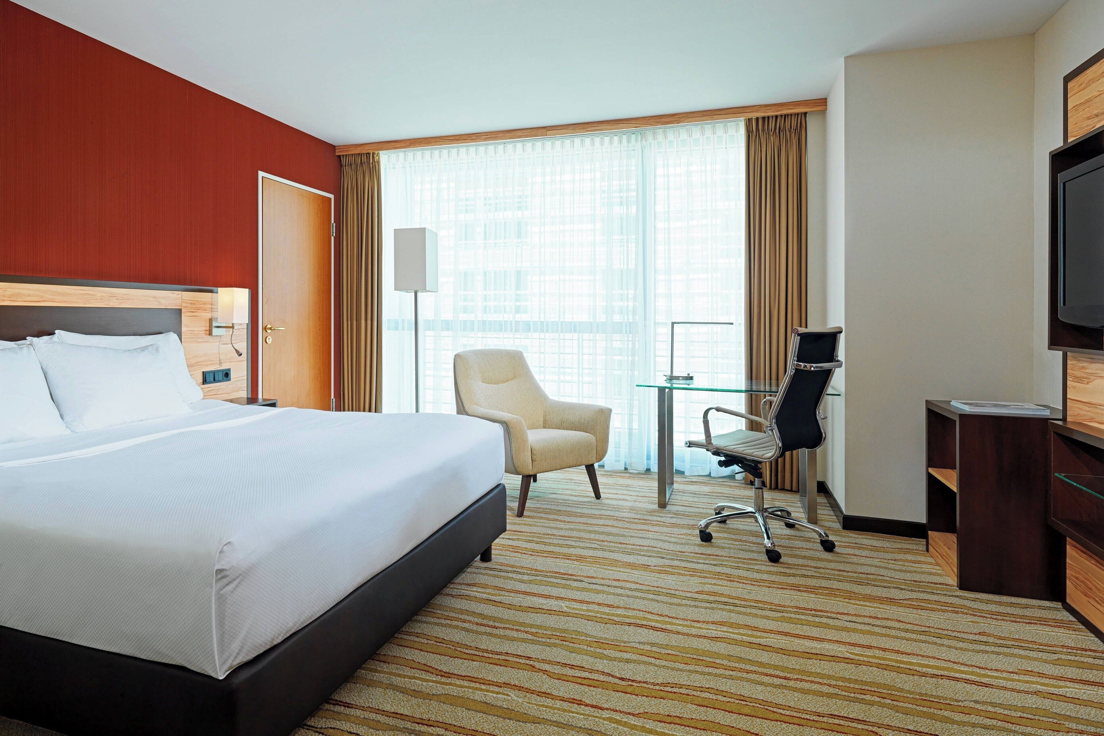 Een hotelkamer met een groot bed, een bureau met stoel, een televisietoestel en een groot raam met gordijnen.