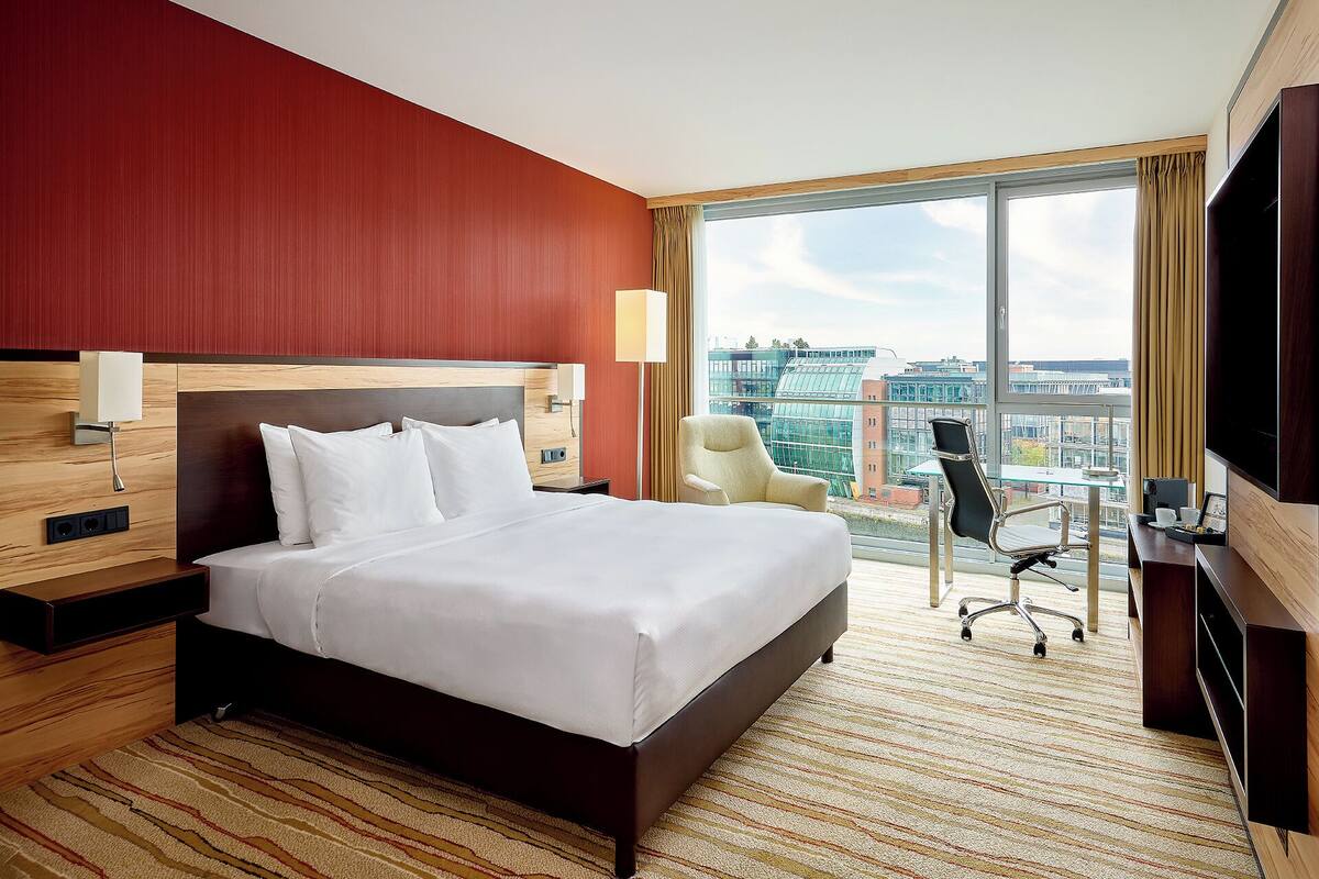 Habitación de hotel con una cama grande, un escritorio con una silla, un televisor y vistas a la ciudad a través de amplias ventanas.