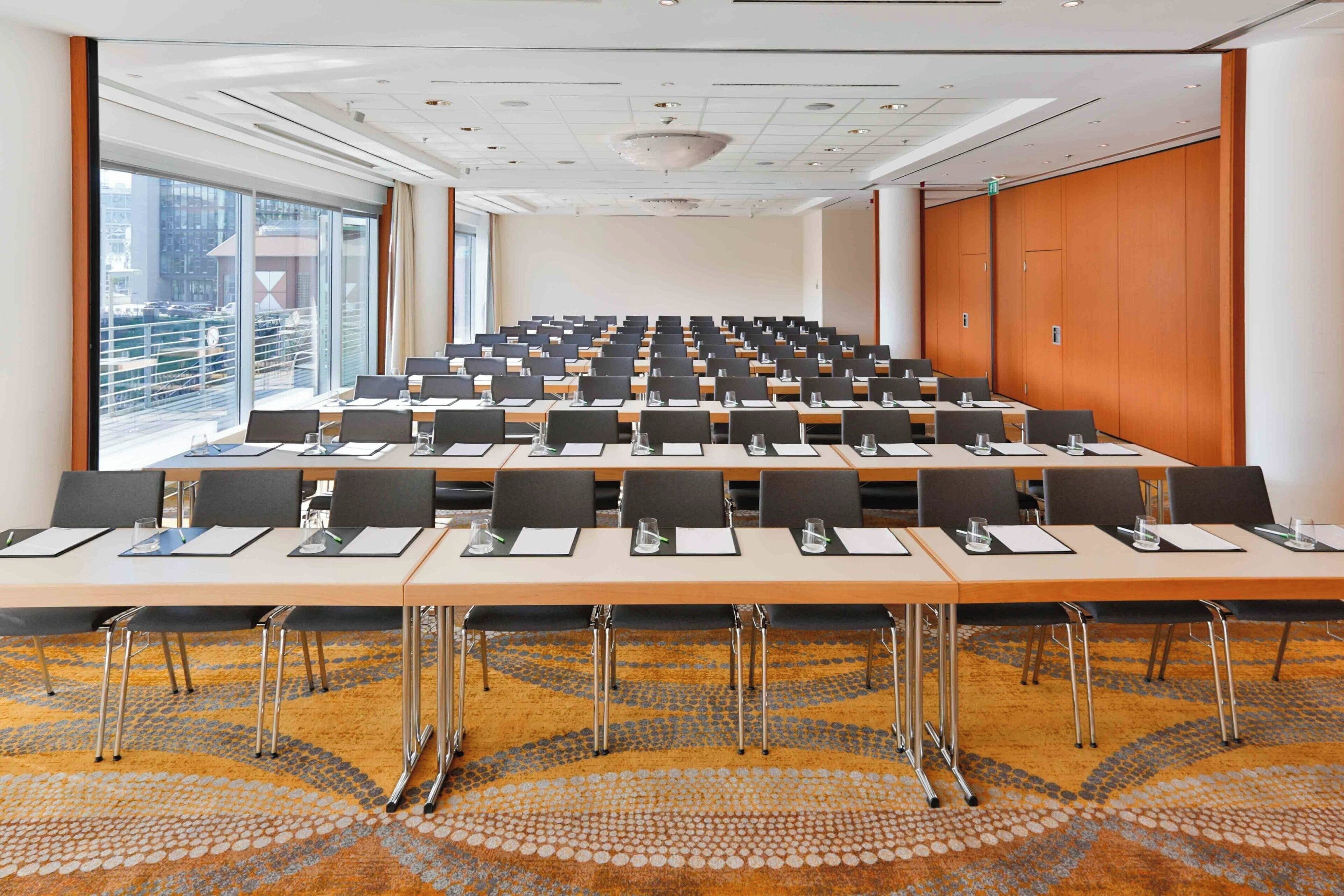 A conference room with multiple tables and chairs, large windows with a city view, and a patterned carpet.