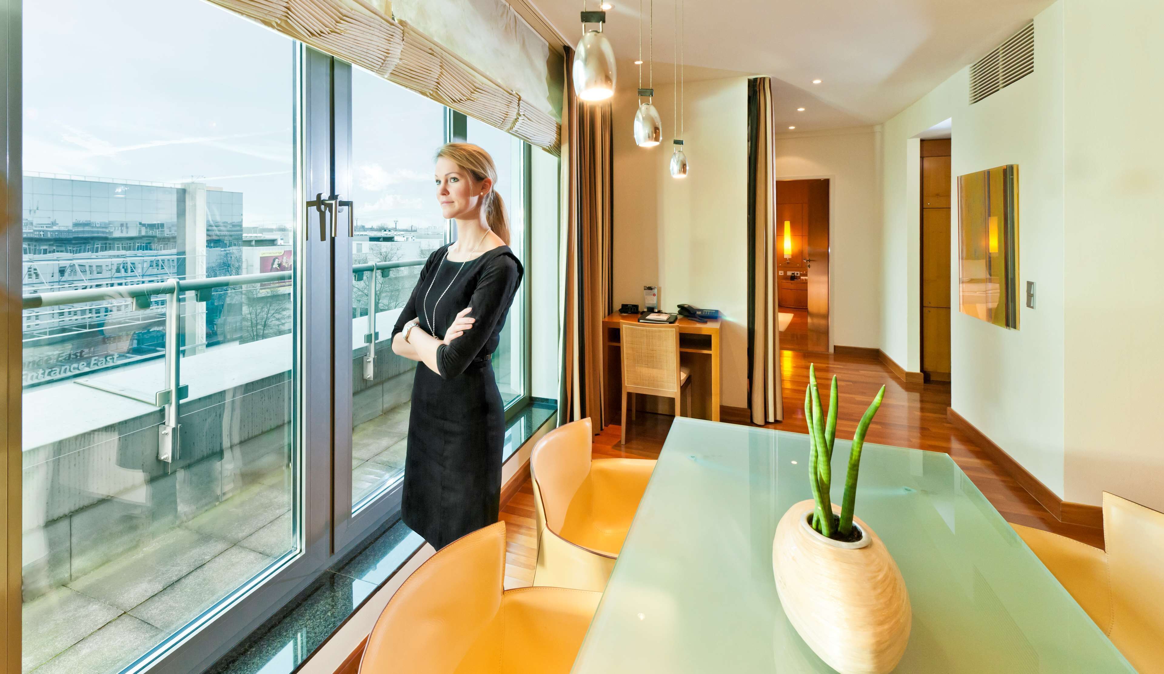 Ein modernes Hotelzimmer mit großem Fenster und Blick auf die Stadt, einem Glastisch mit Topfpflanze sowie einer Sitzecke mit orangefarbenen Stühlen.