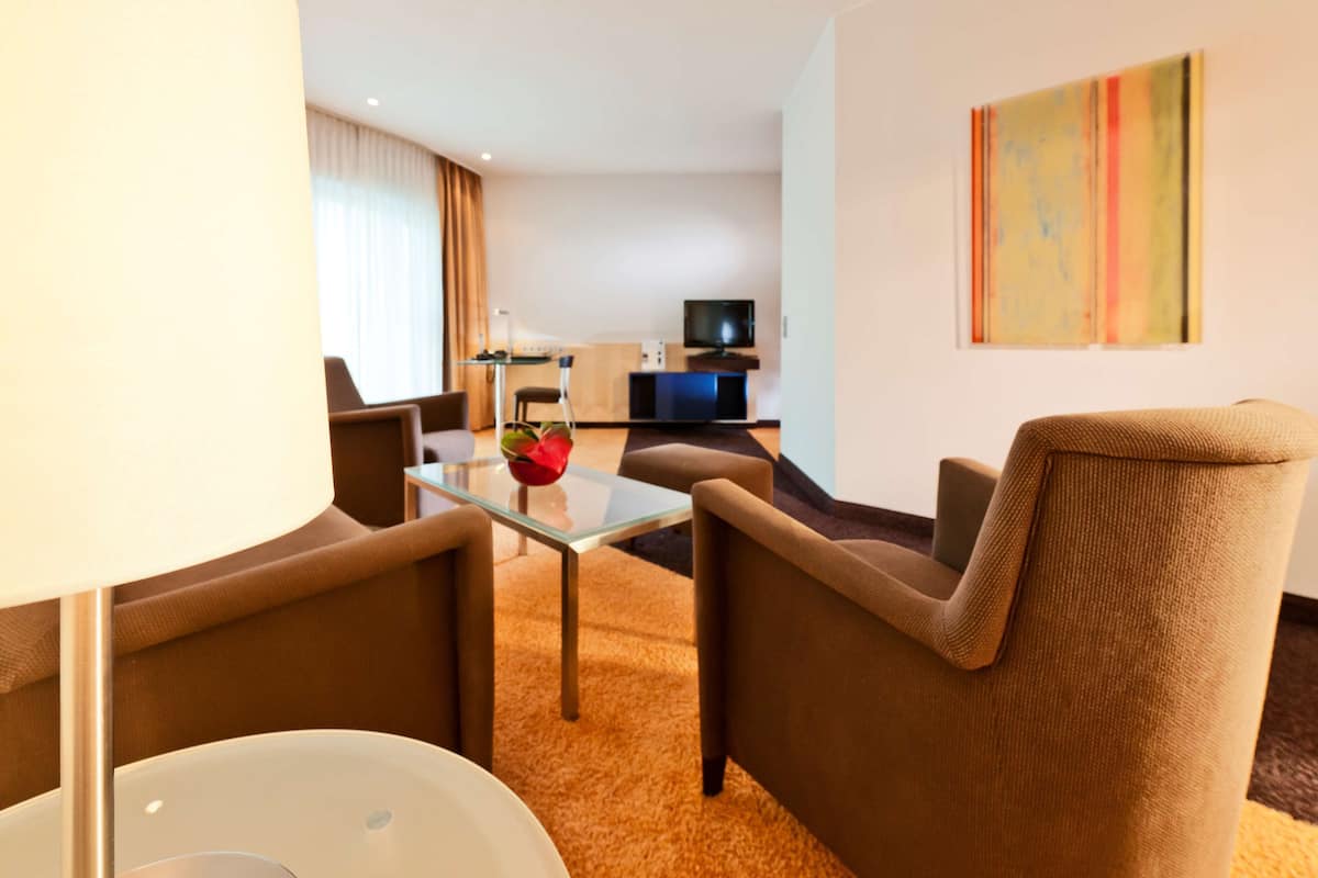 Ein modernes Hotelzimmer mit Wohnbereich, ausgestattet mit einem Glastisch, bequemen braunen Sesseln, einem Flachbildfernseher und einem farbenfrohen, abstrakten Gemälde an der Wand.