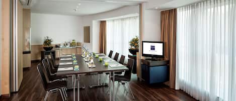 A conference room with a long table, chairs, a TV, and a kitchen area in the background.