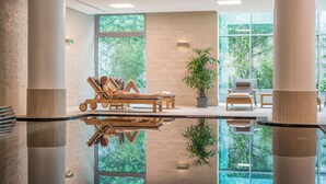 Un espace piscine intérieur moderne, doté de chaises longues et d’un plancher en verre qui reflète la verdure environnante.
