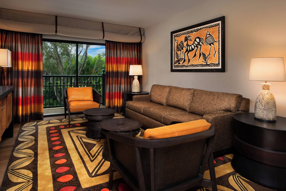 A hotel room with a sofa, armchair, coffee table, and a dining table. There is a window with curtains, a painting on the wall, and a lamp on the side table.