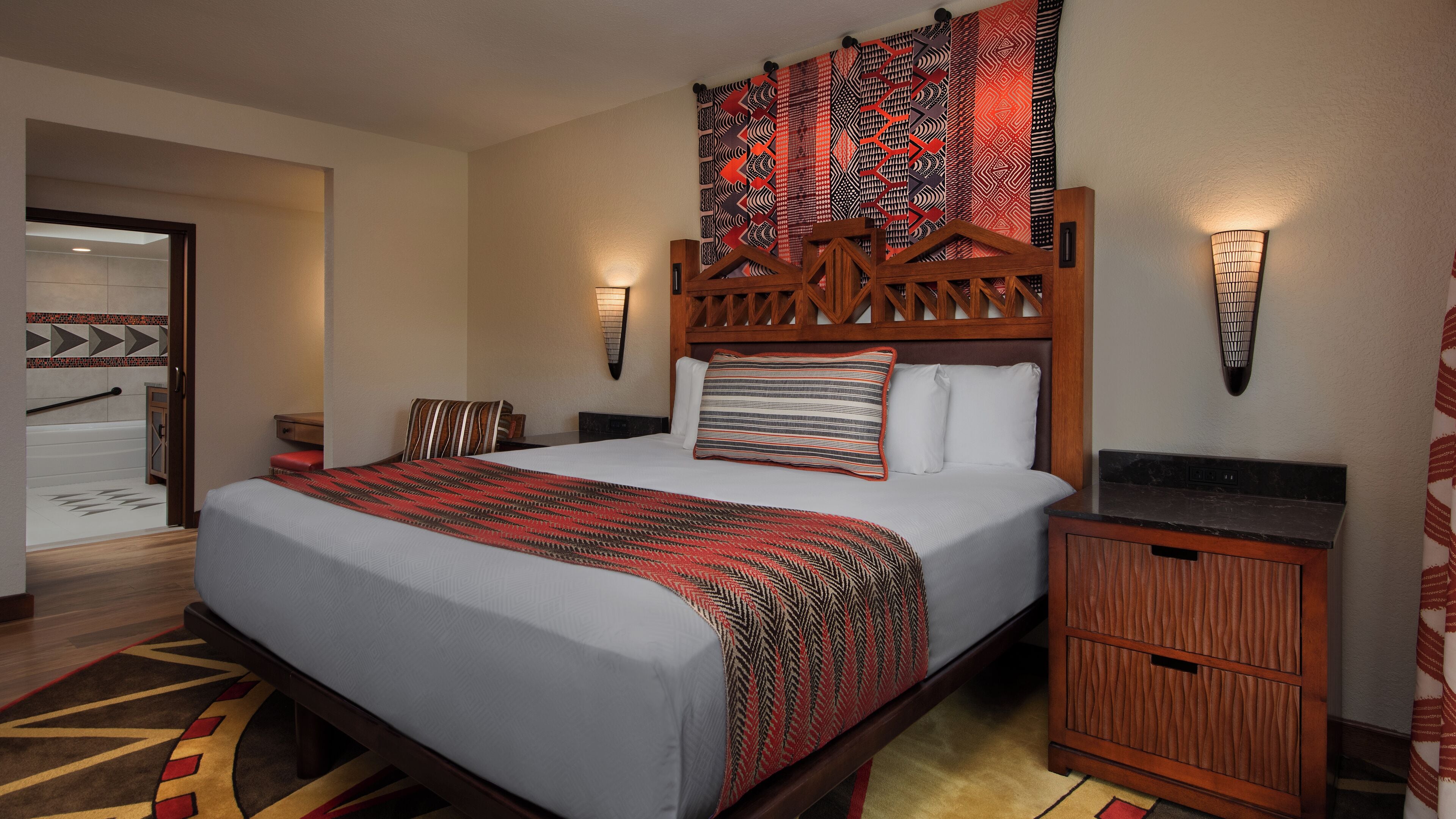 A hotel room with a large bed, wooden headboard, two nightstands, a wall-mounted lamp, and a patterned tapestry.