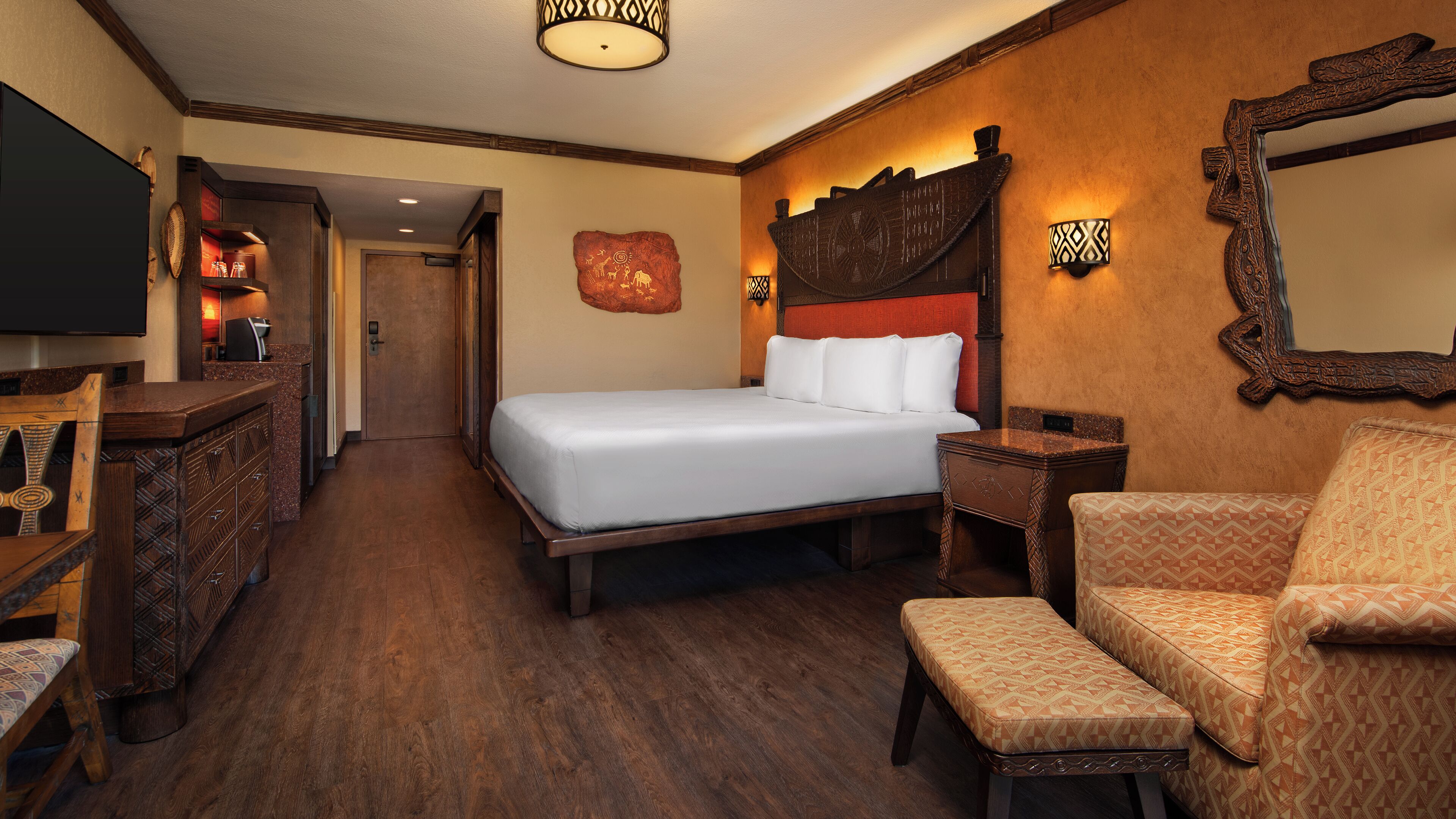 A hotel room with a large bed, a wooden dresser, a TV, a seating area with a patterned sofa, and a mirror on the wall.