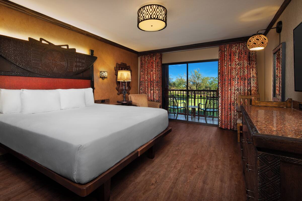 A hotel room with a large bed, a wooden headboard, a nightstand, a lamp, and a view of a golf course through the window.