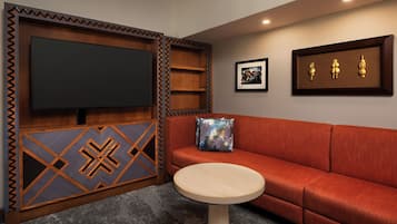 A room with a large flat-screen TV mounted on a wooden panel, a red sofa with a patterned cushion, a small round wooden table, and framed artwork on the wall.