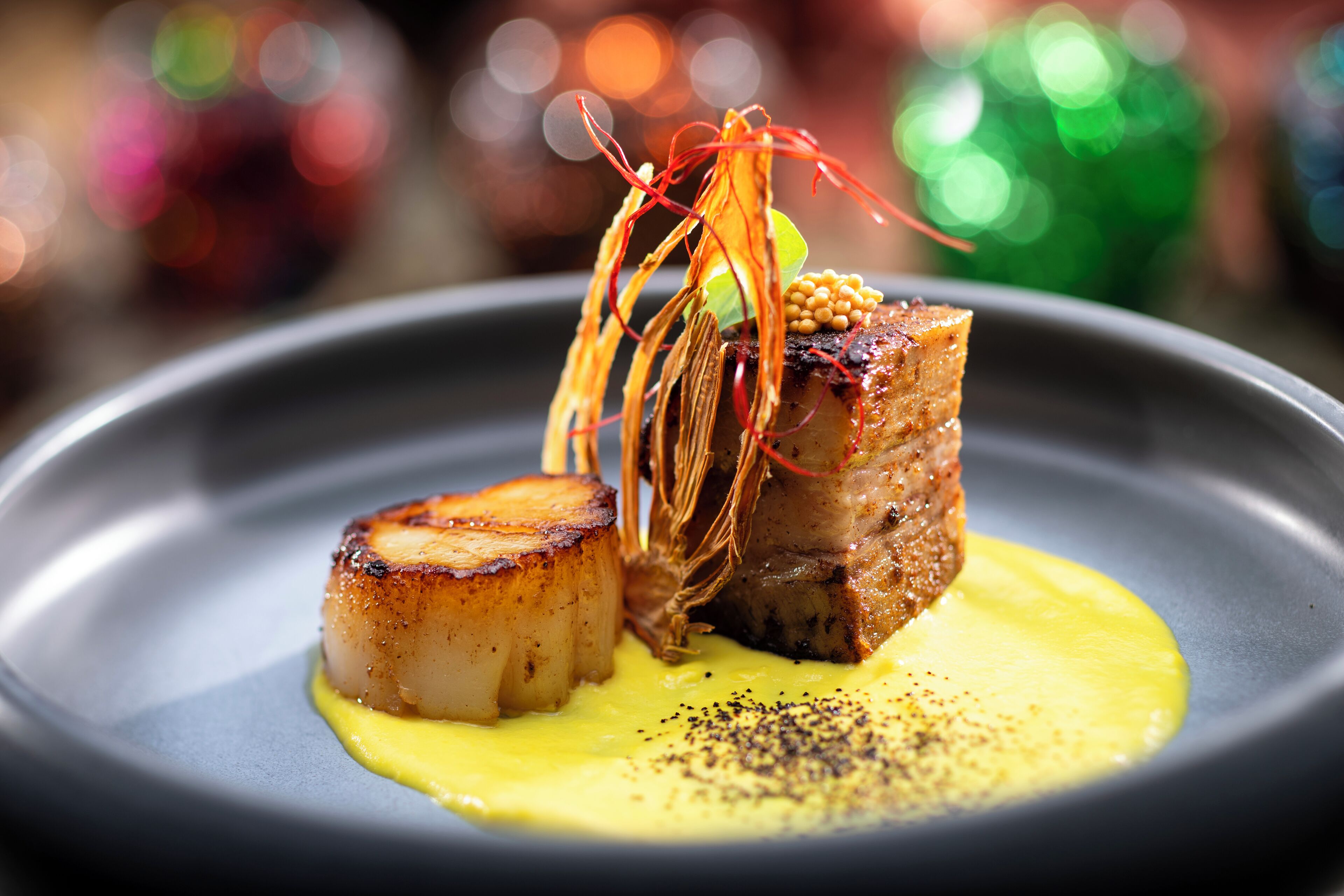 A plated dish with seared scallops and a piece of meat, garnished with a yellow sauce and black pepper.