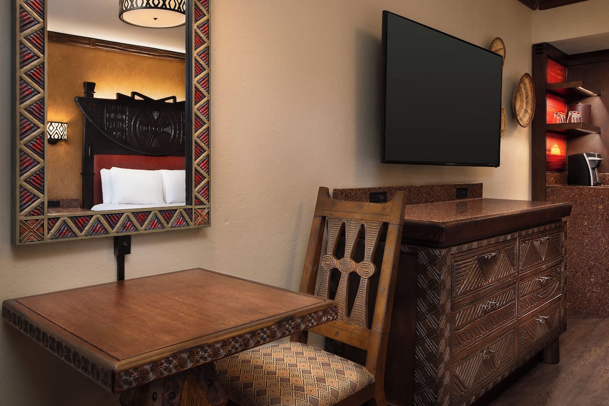 A hotel room with a wooden dining table, a patterned chair, a large mirror with a decorative frame, a flat-screen TV mounted on the wall, and a wooden cabinet with drawers.