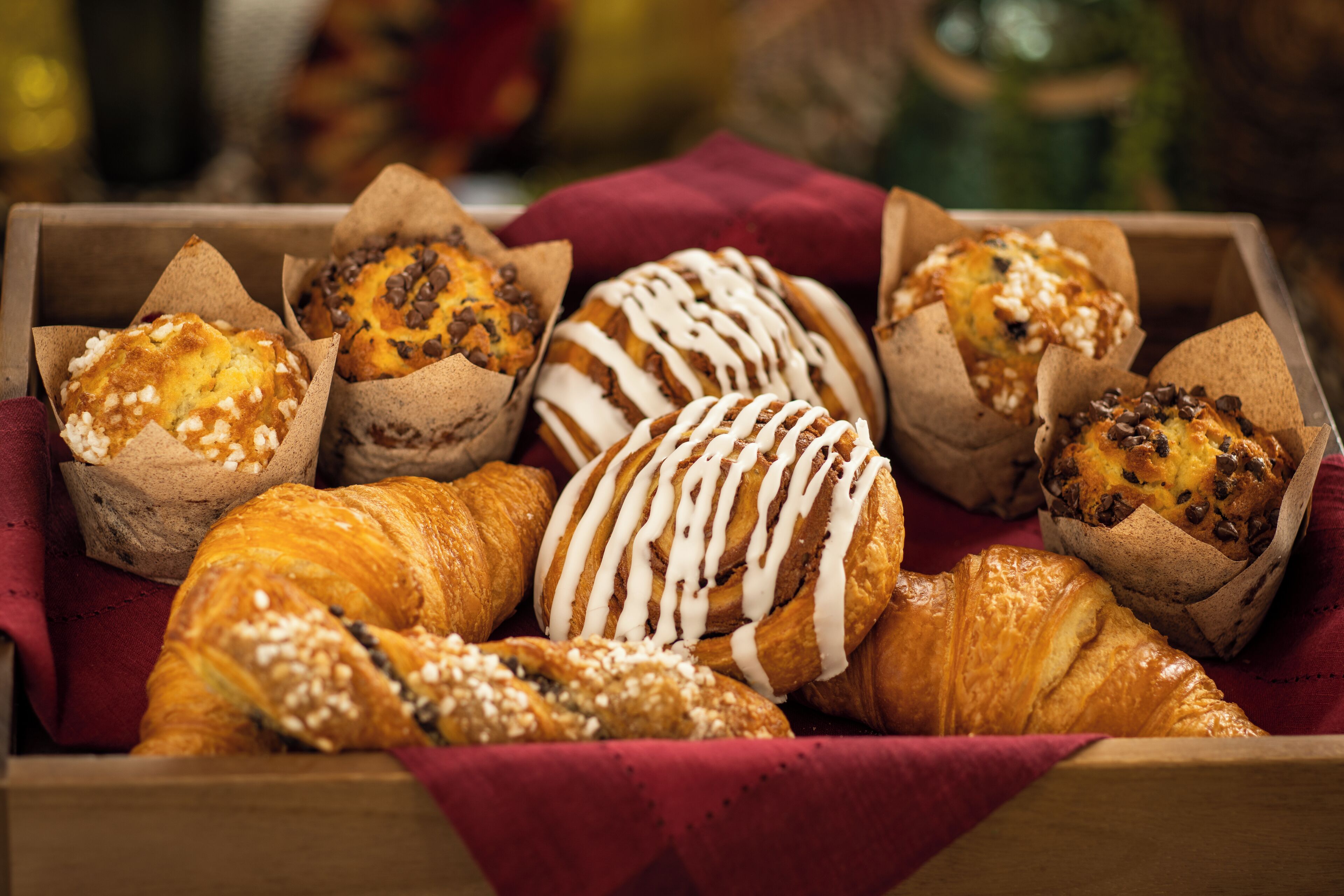 A wooden tray with various baked goods, including muffins and a croissant, arranged on a red cloth.