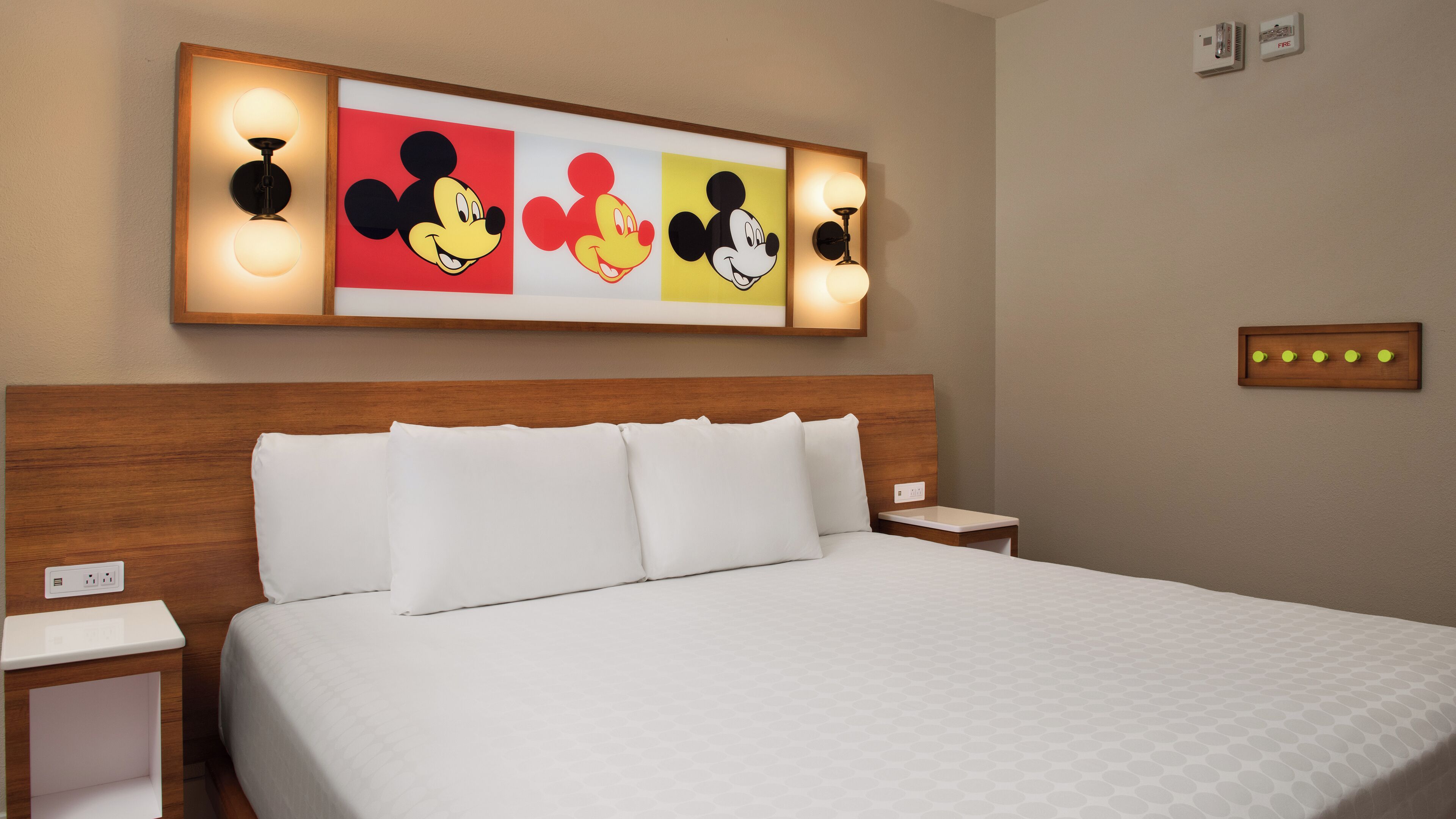 A hotel room with a large bed, white bedding, a wooden headboard, and wall-mounted artwork featuring Mickey Mouse.
