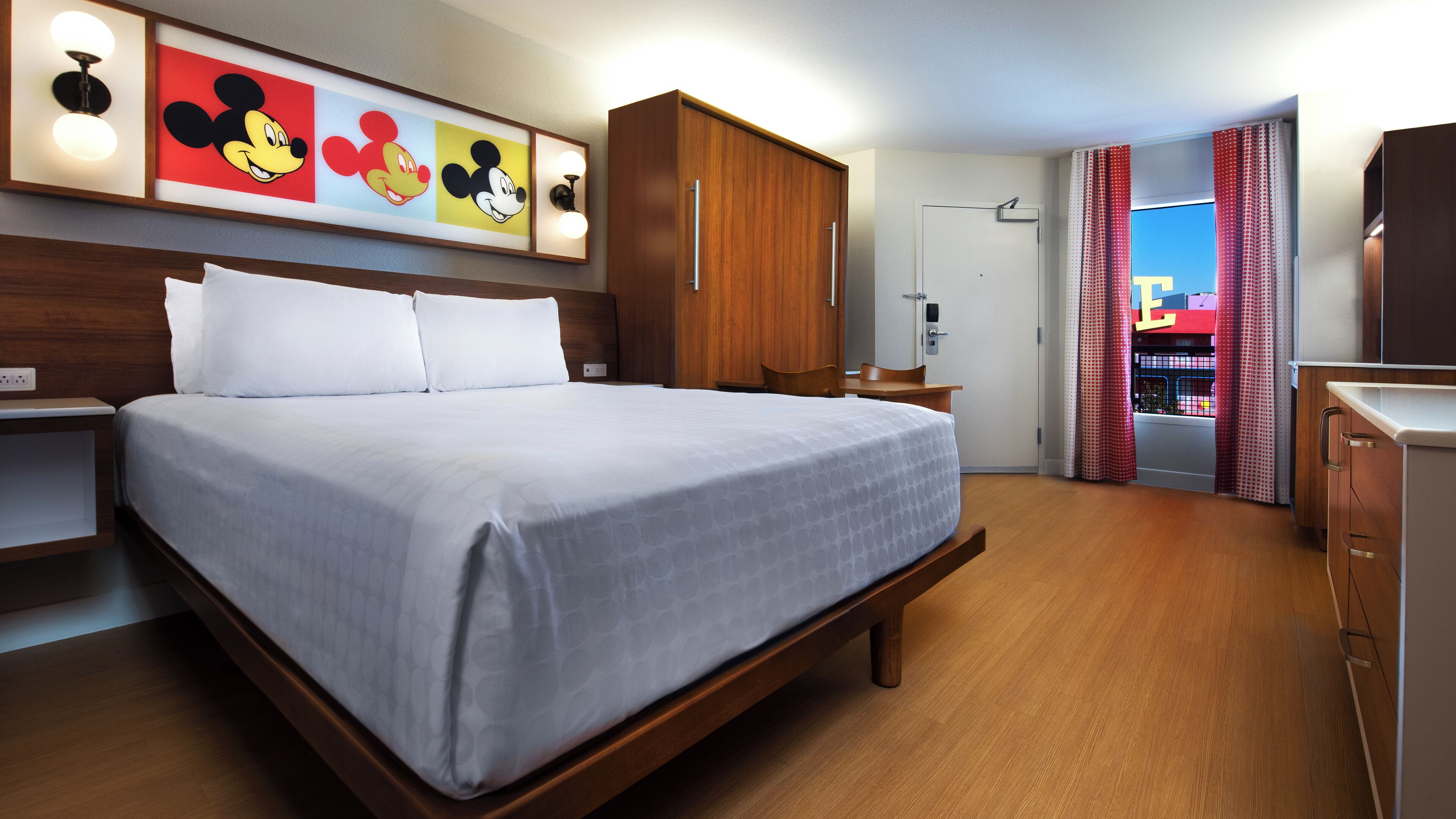 Ein Hotelzimmer mit einem großen Bett, einem Holzkopfstück, einem Schreibtisch, einem Stuhl und einem Kleiderschrank. An der Wand befinden sich Bilder von Mickey Mouse.