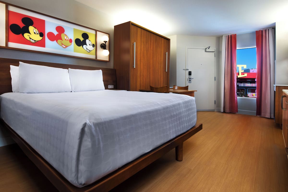 A hotel room with a large bed, wooden headboard, a desk, a chair, and a wardrobe. There are Mickey Mouse paintings on the wall.