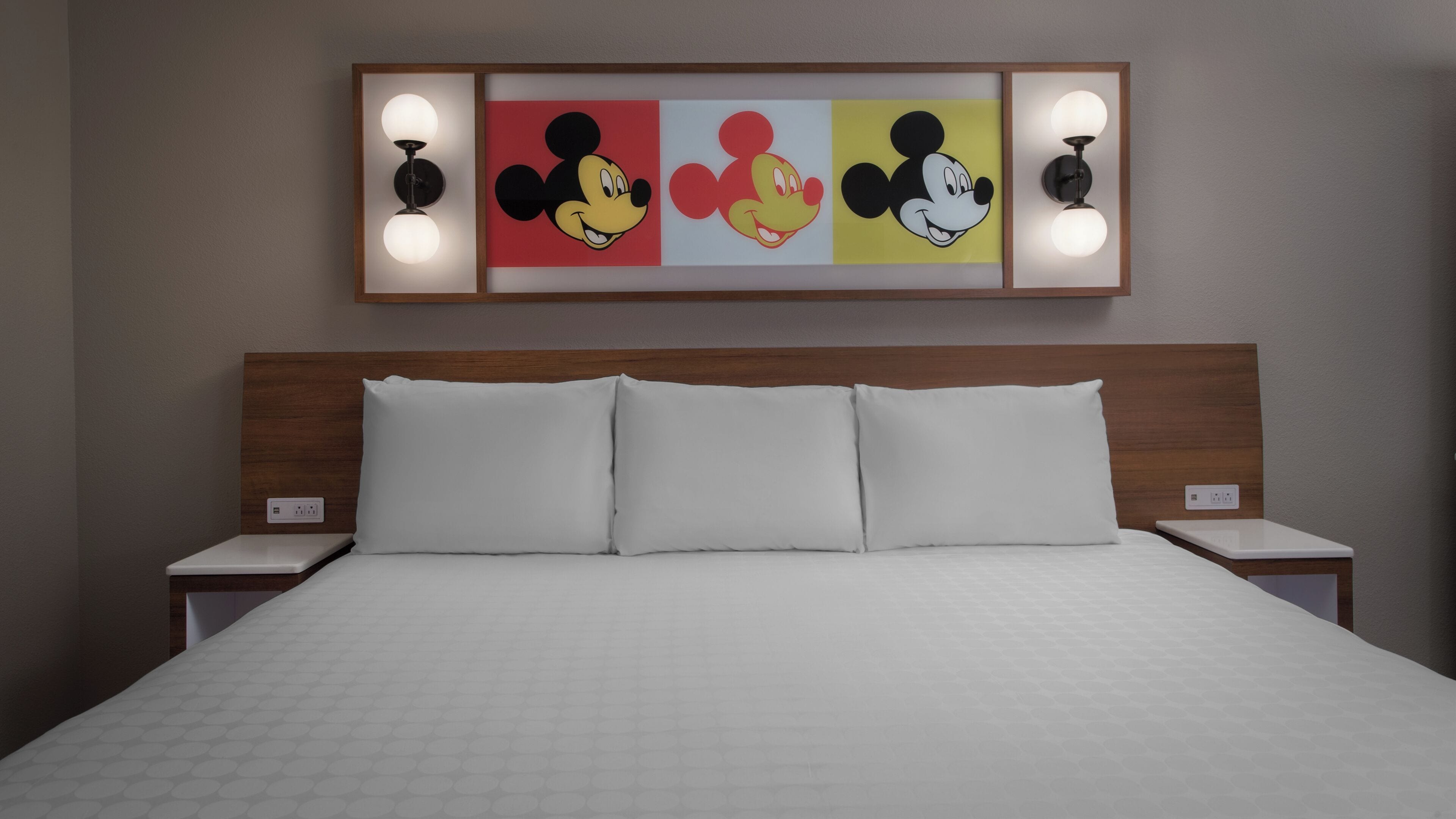 A hotel room with a bed, white bedding, a headboard, and three framed pictures of Mickey Mouse on the wall.