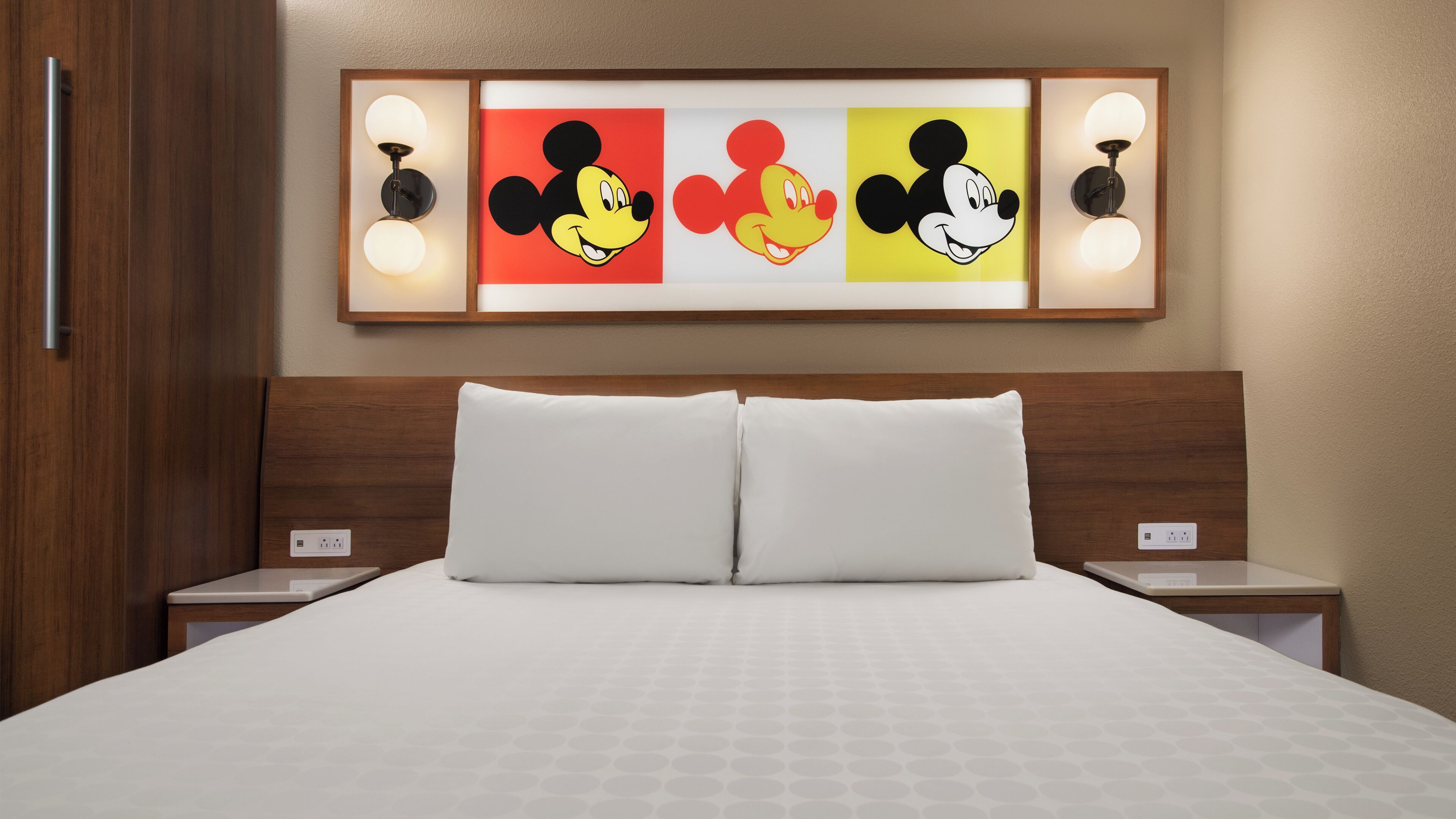 Ein Hotelzimmer mit einem großen Bett, zwei Nachttischen und einer Wand mit gerahmten Bildern von Mickey Mouse.