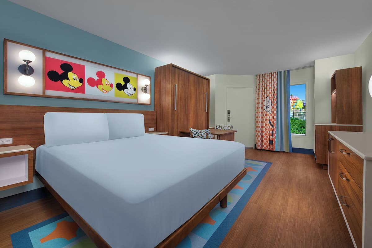 A hotel room with a large bed, wooden wardrobe, a desk with a chair, and Mickey Mouse artwork on the wall.