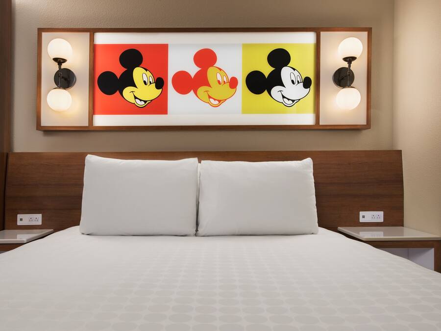 A hotel room with a large bed, two bedside tables, and a wall-mounted artwork featuring Mickey Mouse.