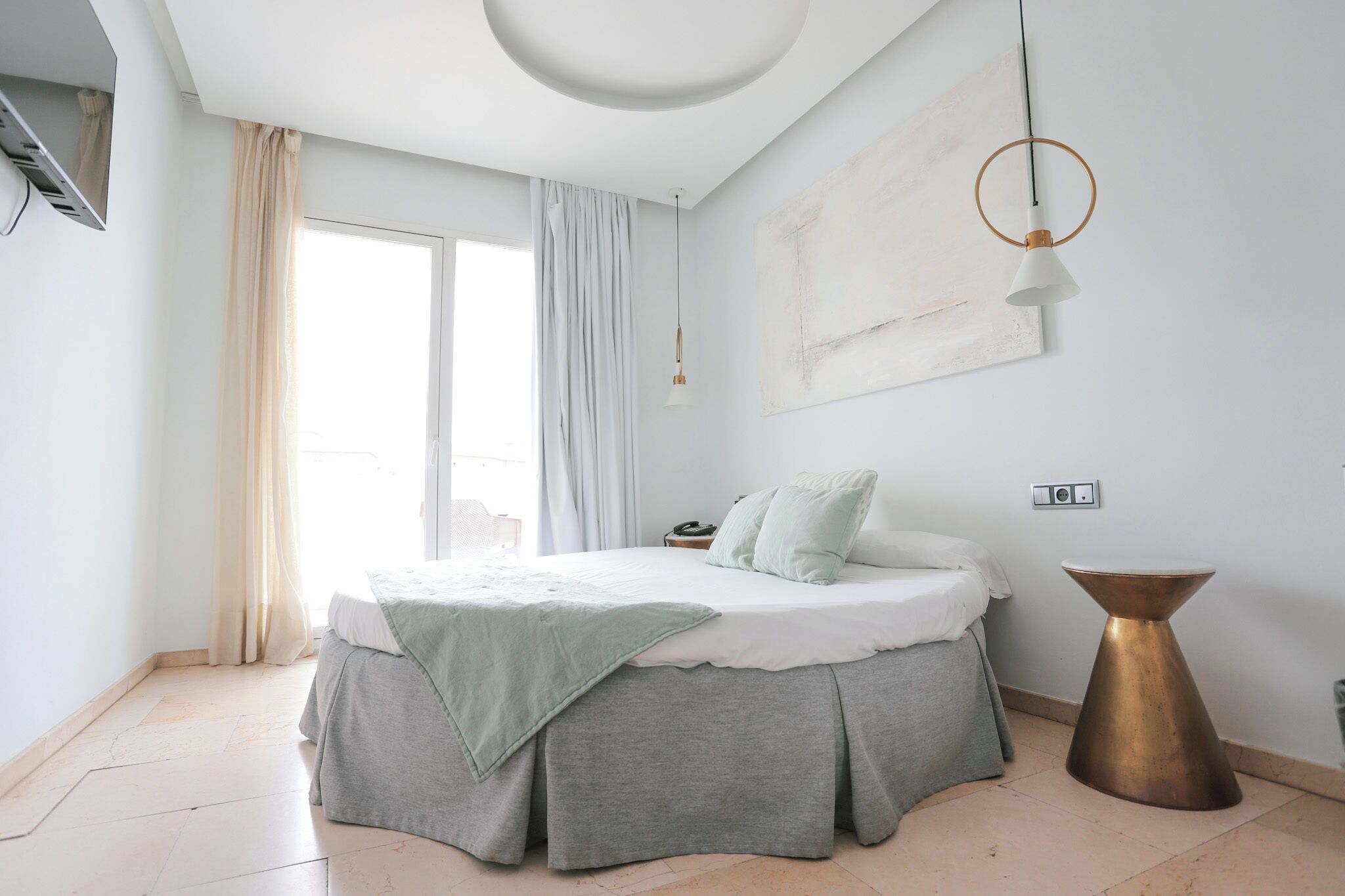 Une chambre à coucher comprenant un lit, des rideaux, un luminaire suspendu, une table de chevet et une œuvre d’art murale.
