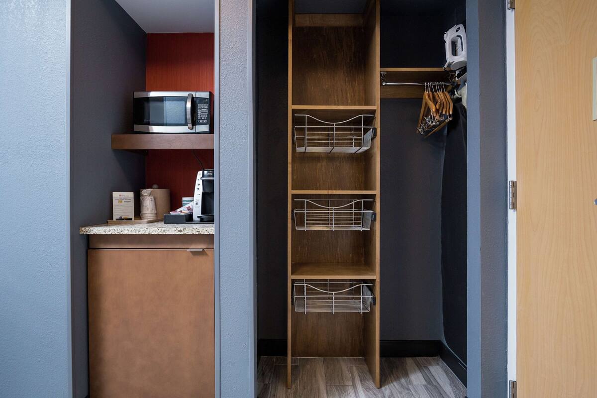 A compact kitchen with a microwave, a coffee machine, and a wooden shelf with wire baskets.