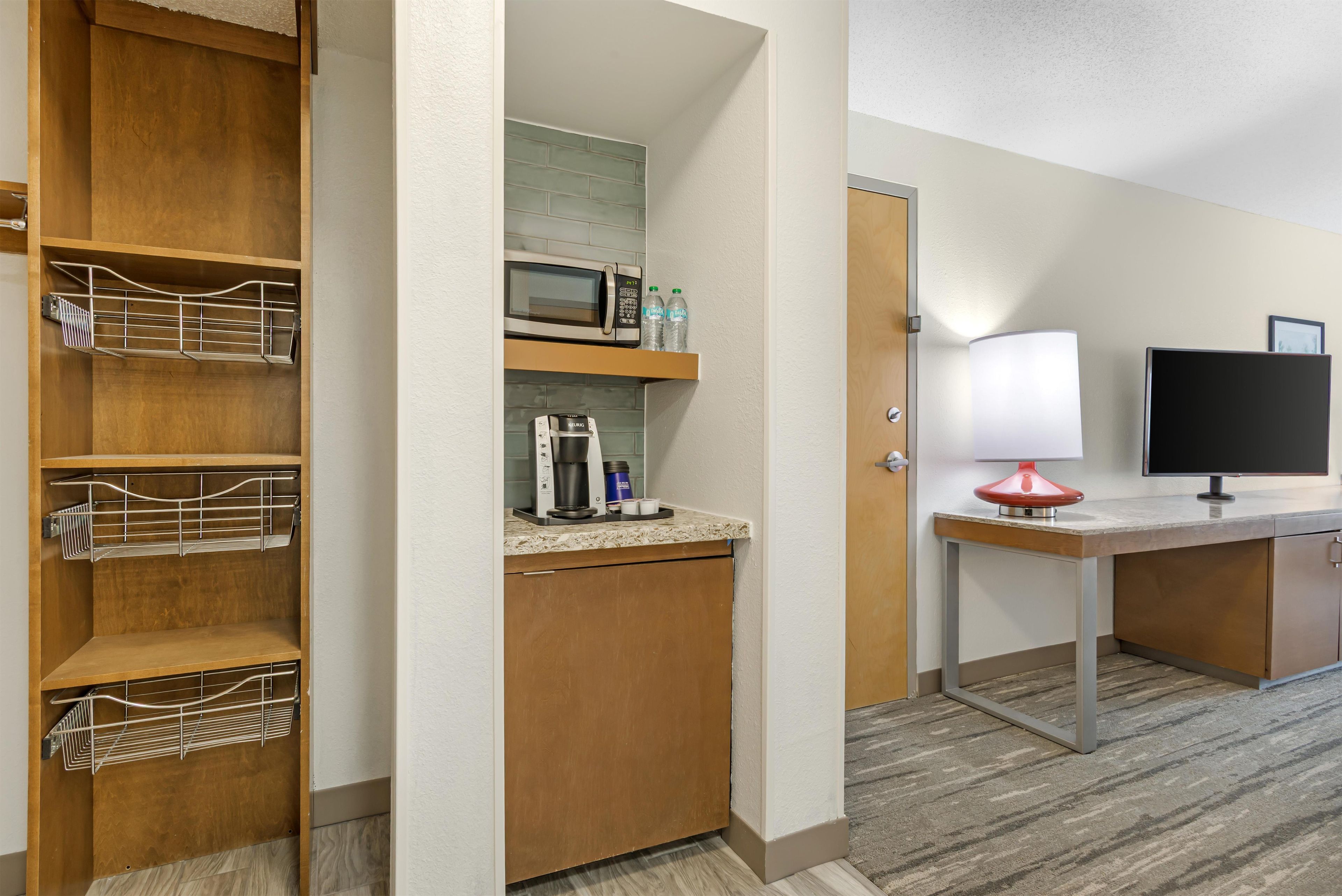 A hotel room with a wooden wardrobe, a microwave, a coffee machine, a small table with a lamp, and a television.