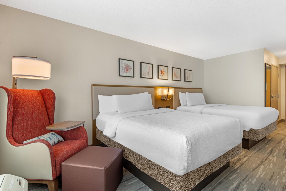 A hotel room with two beds, a desk, a chair, and a lamp.