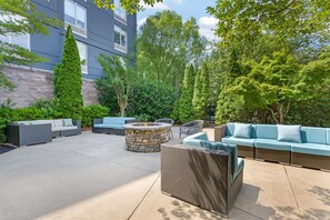 An outdoor patio area with a stone fire pit, wicker furniture, and a view of a modern building. - Hilton Garden Inn Atlanta Northpoint (Alpharetta)