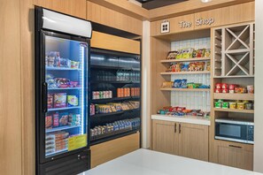 A snack and beverage refrigerator stocked with various items, a section labeled 'The Shop' with snacks and canned goods, and a microwave oven. - Hilton Garden Inn Atlanta Northpoint (Alpharetta)