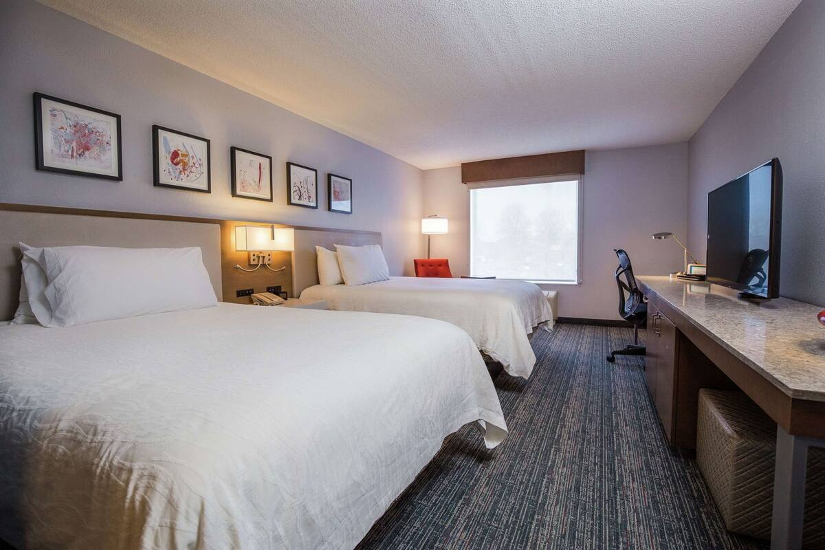 Quarto de hotel com duas camas, uma escrivaninha, televisão e quadros nas paredes.