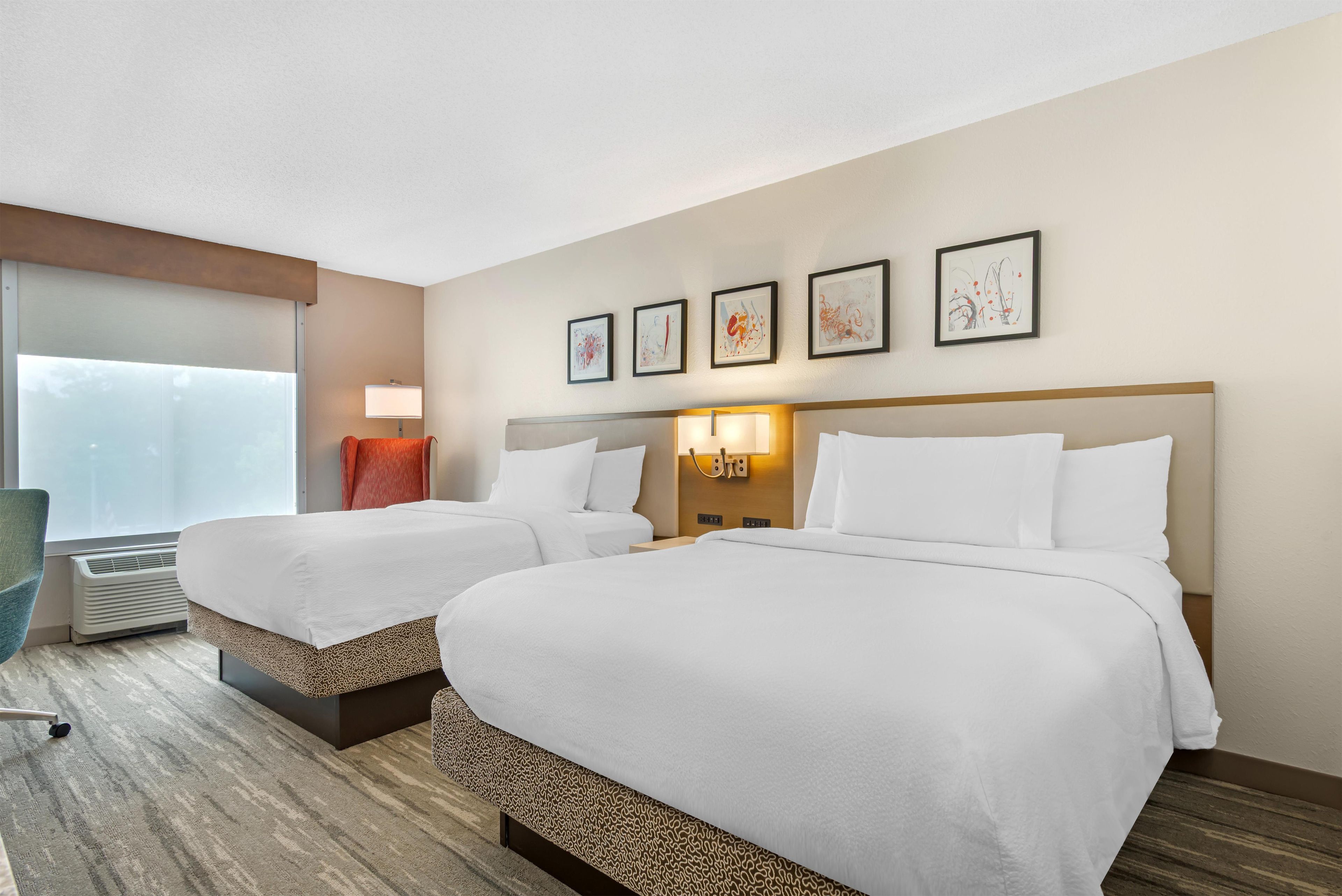 A hotel room with two beds, a window with curtains, a desk with a chair, and artwork on the wall.