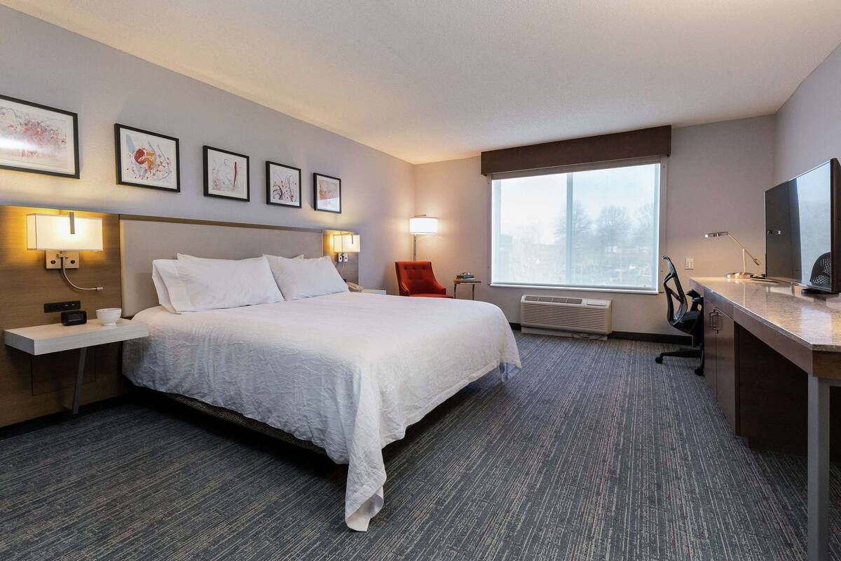 A hotel room with a bed, bedside tables, a desk, a chair, a window, and artwork on the wall.