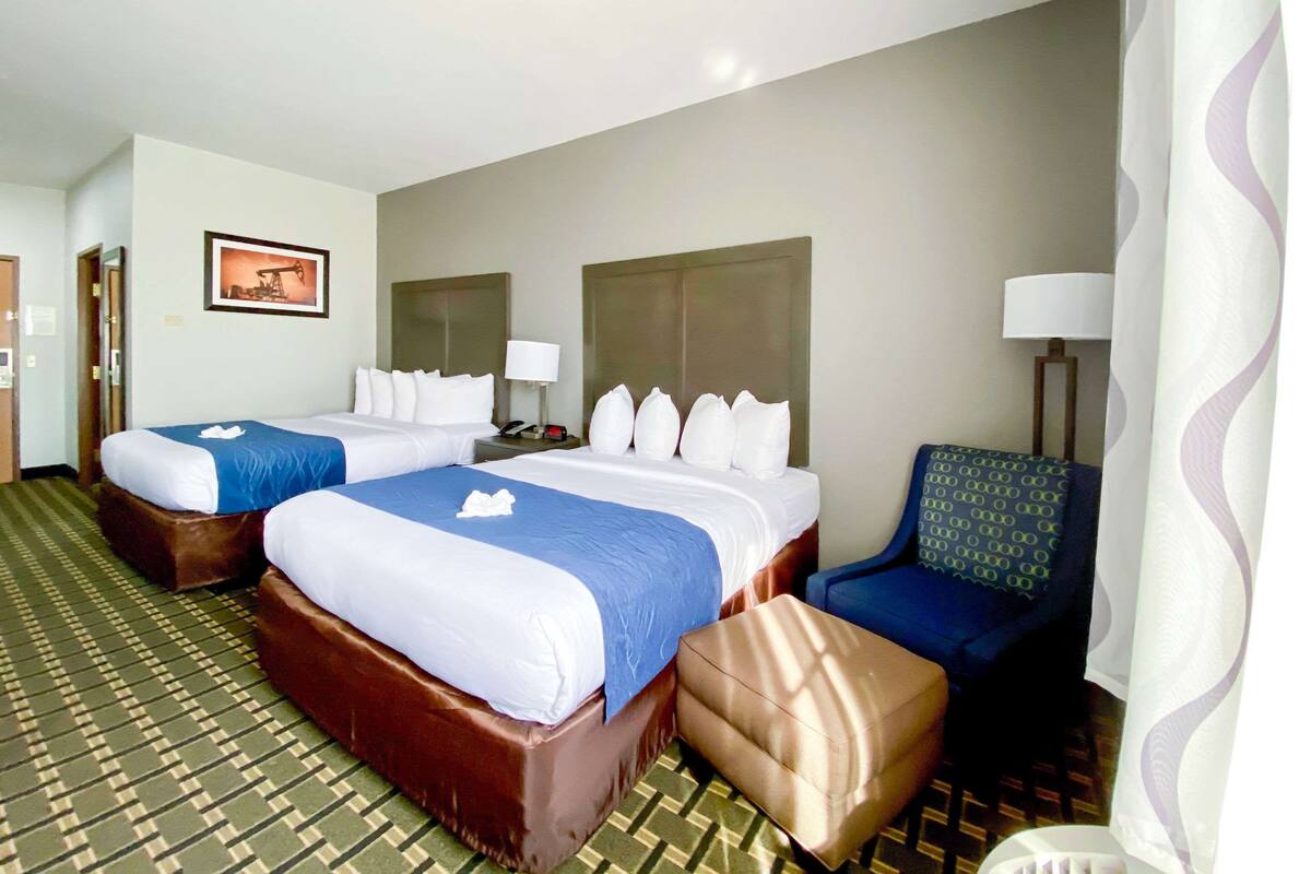 A hotel room with two beds, a blue chair, a brown ottoman, a lamp, and a picture on the wall.