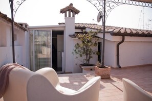 Une terrasse avec deux chaises blanches, une plante en pot et une porte vitrée donnant sur une pièce.