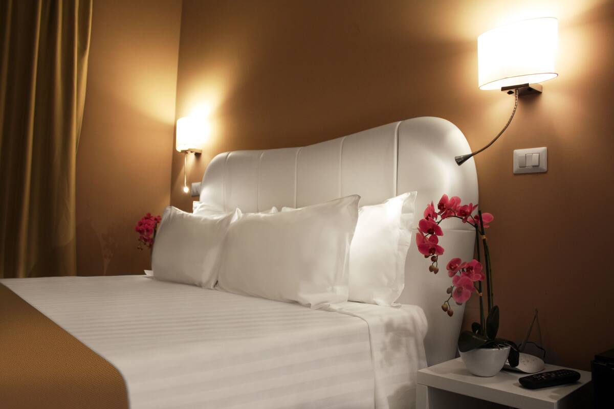 A hotel room with a bed, white headboard, two bedside lamps, a nightstand with a remote control, and a potted orchid.