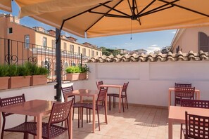 Une terrasse extérieure avec des tables et des chaises, un grand parasol et une vue sur les bâtiments en arrière-plan.