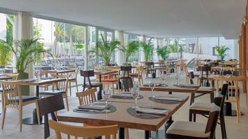 Un restaurante espacioso con varias mesas y sillas, amplias ventanas que ofrecen vistas al exterior y plantas en macetas que aportan verdor.