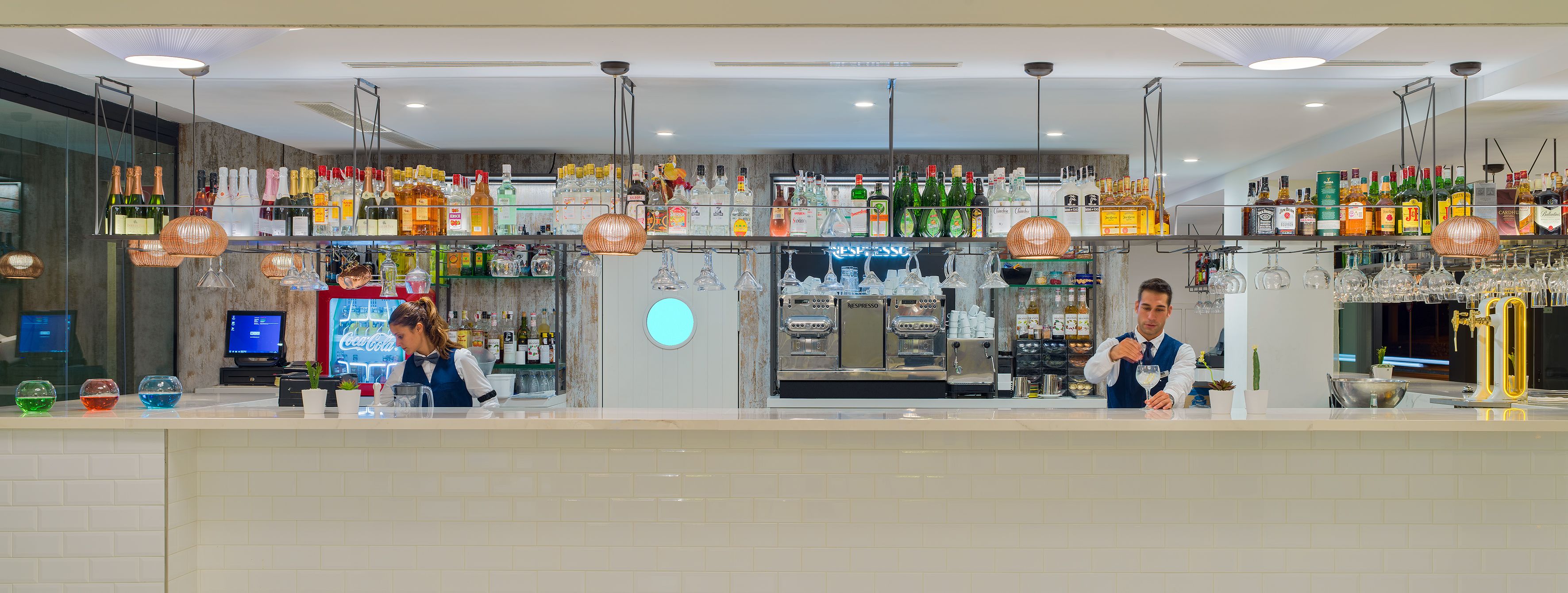 A bar with multiple shelves of liquor bottles and glasses, a coffee machine, and two bartenders at work.