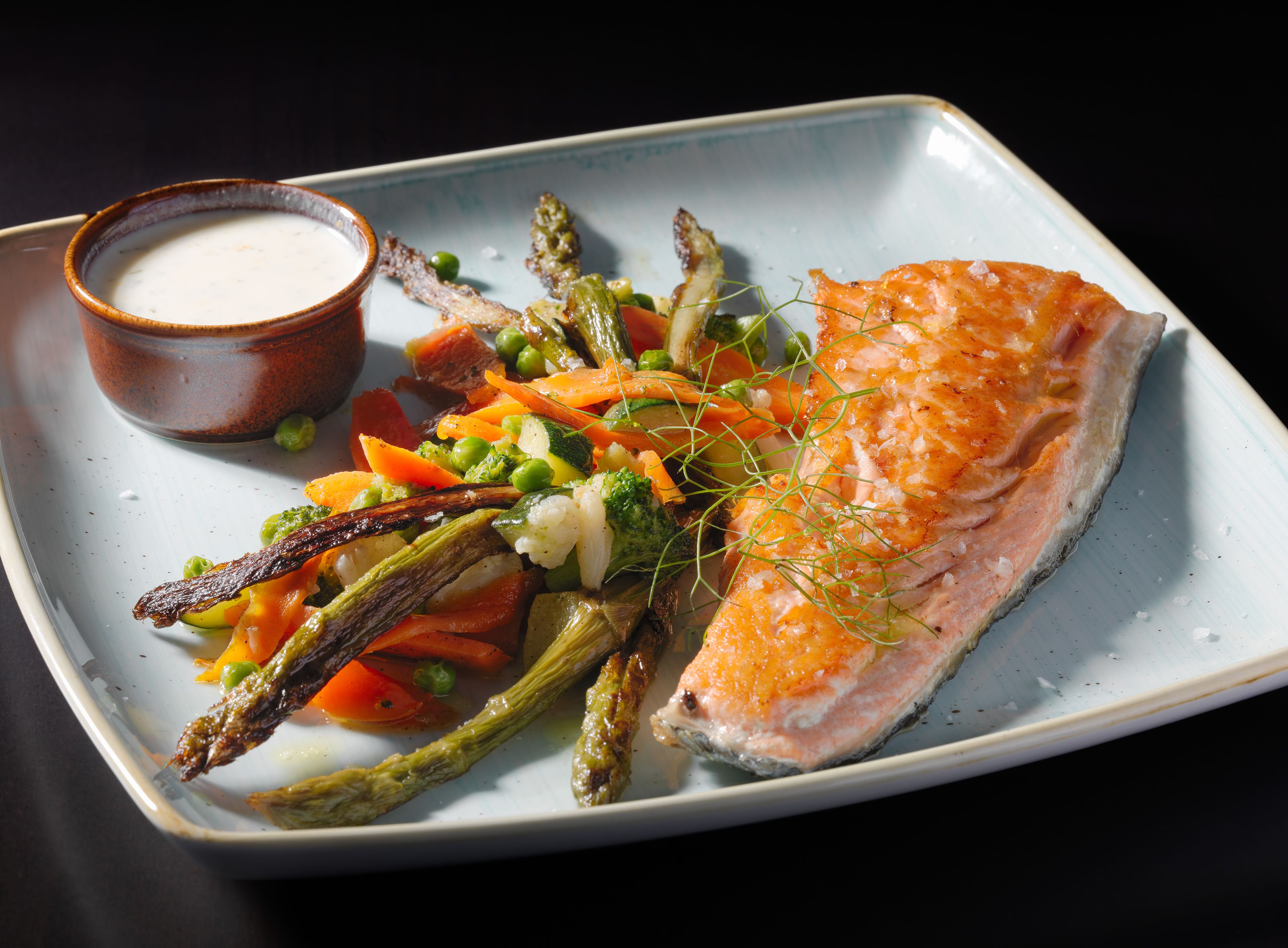 A plate with salmon, asparagus, peas, carrots, and a small bowl of sauce.